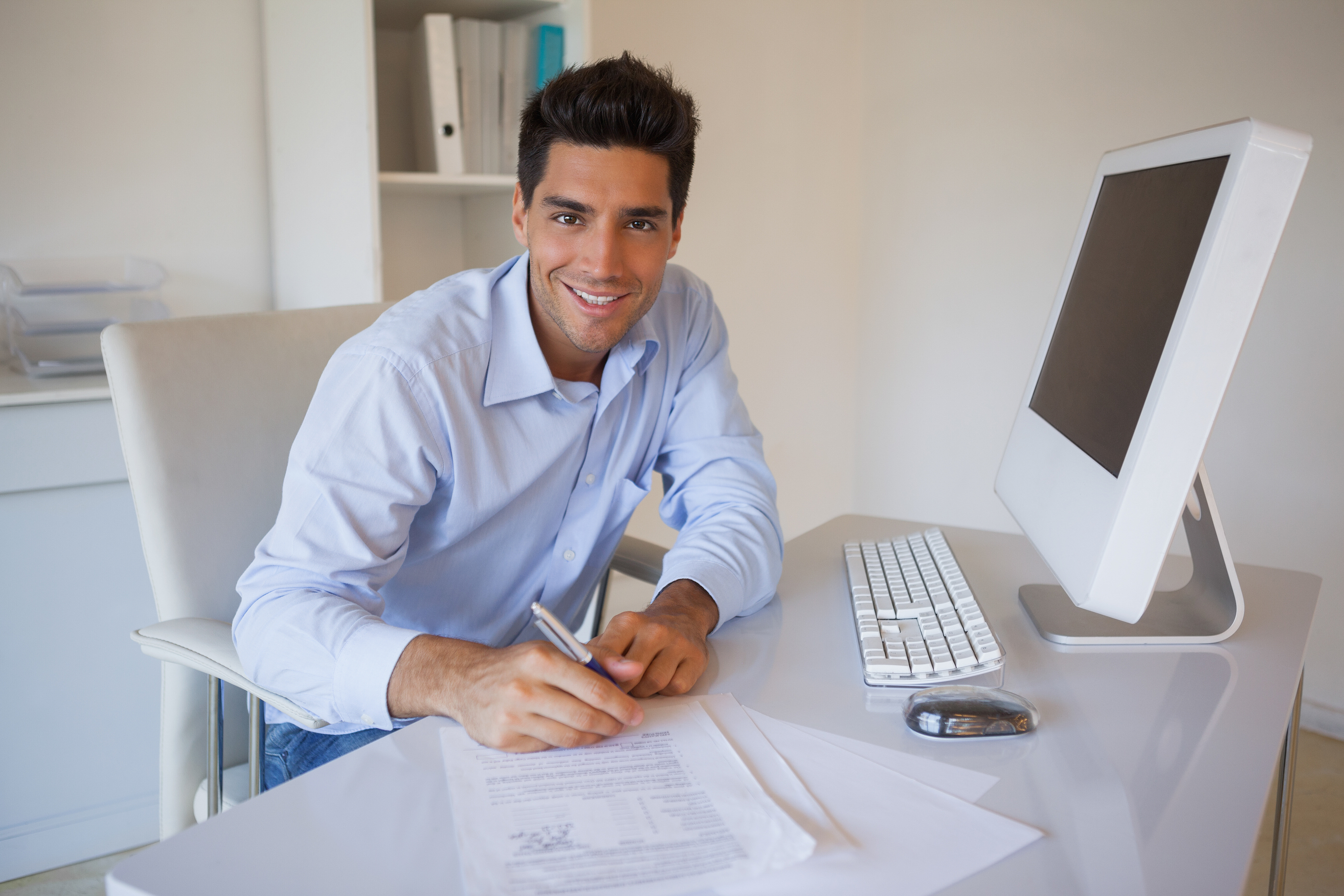 A man wearing a blue shirt is sitting at a desk in an office. He has one hand resting on a document, the other holding a pen. He is looking at the camera and smiling.